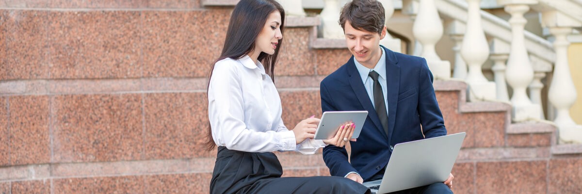 Business man and woman collaborating on a tablet and laptop in front of a government building